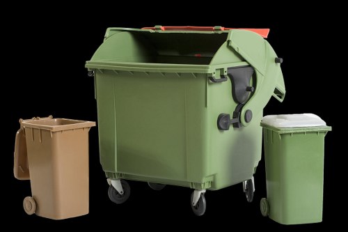 The image displays three different wheelie bins positioned against a black background, highlighting a scene typical for waste disposal or rubbish removal. The largest bin, centrally located, is a standard green plastic wheelie bin with a textured surface, black handles on each side, and twin black wheels at the base, suggesting it is used for general waste collection. To the left, there is a smaller beige-colored bin made of plastic, featuring a flat lid and two small wheels at its base, likely intended for household waste. To the right, a slightly taller green plastic bin with a matching lid is visible, also on small wheels, typically used for garden or recyclable waste. The lighting emphasizes the textures and colours of the bins, with reflections on their surfaces indicating a well-lit environment, possibly outdoors or in a storage area. The scene is related to waste management services, such as those offered by Commercial Waste Woking, serving the Woking area for rubbish collection and disposal needs based in Surrey, within the postcode region prefixed by GU. The arrangement of bins suggests readiness for collection or storage in a commercial or residential setting, reflecting typical waste disposal practices in the UK.