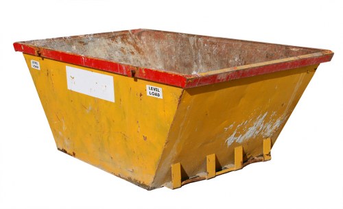 A large industrial skip with a yellow painted exterior and a red frame along the top edge, positioned outdoors on a paved surface. The skip's interior is visible, showing signs of rust and accumulated debris. It features three orange-colored lifting lugs at the front base for hoisting and emptying. The exterior has some paint wear and patches of dirt, indicating regular use. The background is plain and neutral, with no additional objects or environmental context visible. This image reflects typical waste containment containers used in commercial rubbish removal services, such as those offered by Commercial Waste Woking for businesses in Woking and surrounding areas with postcode GU21. The skip's sturdy construction and utilitarian design are characteristic of equipment employed for collecting and transporting waste materials in various commercial and industrial settings, supporting effective rubbish removal operations.